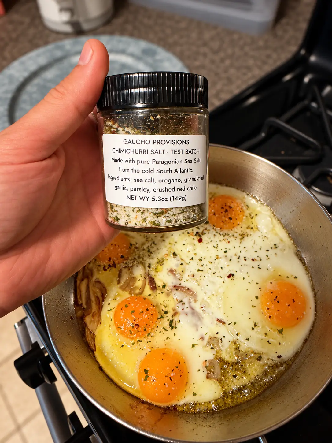 A hand holds a Gaucho Provisions Chimichurri Salt test-batch jar above eggs sprinkled with seasoning in a frying pan.